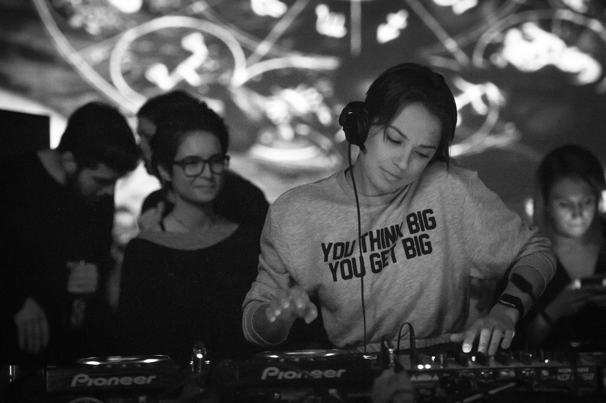 Female DJ wearing headphones behind Pioneer equipment, with guests and projected visuals in the background