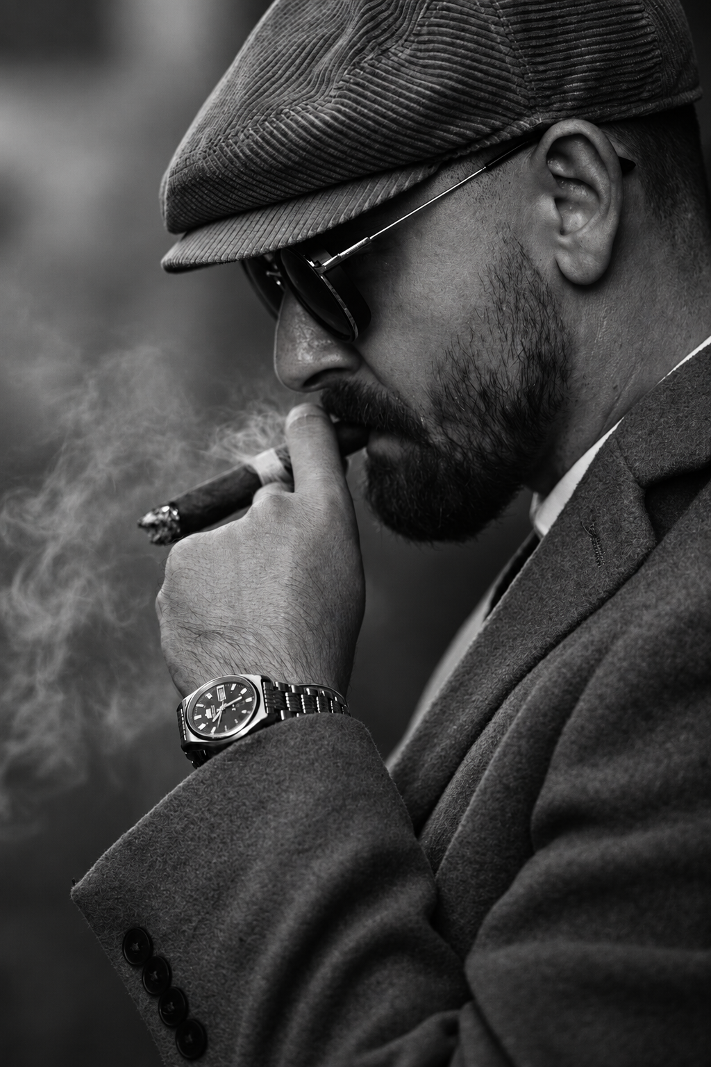 Black-and-white portrait of a man in a flat cap and sunglasses, holding a cigar