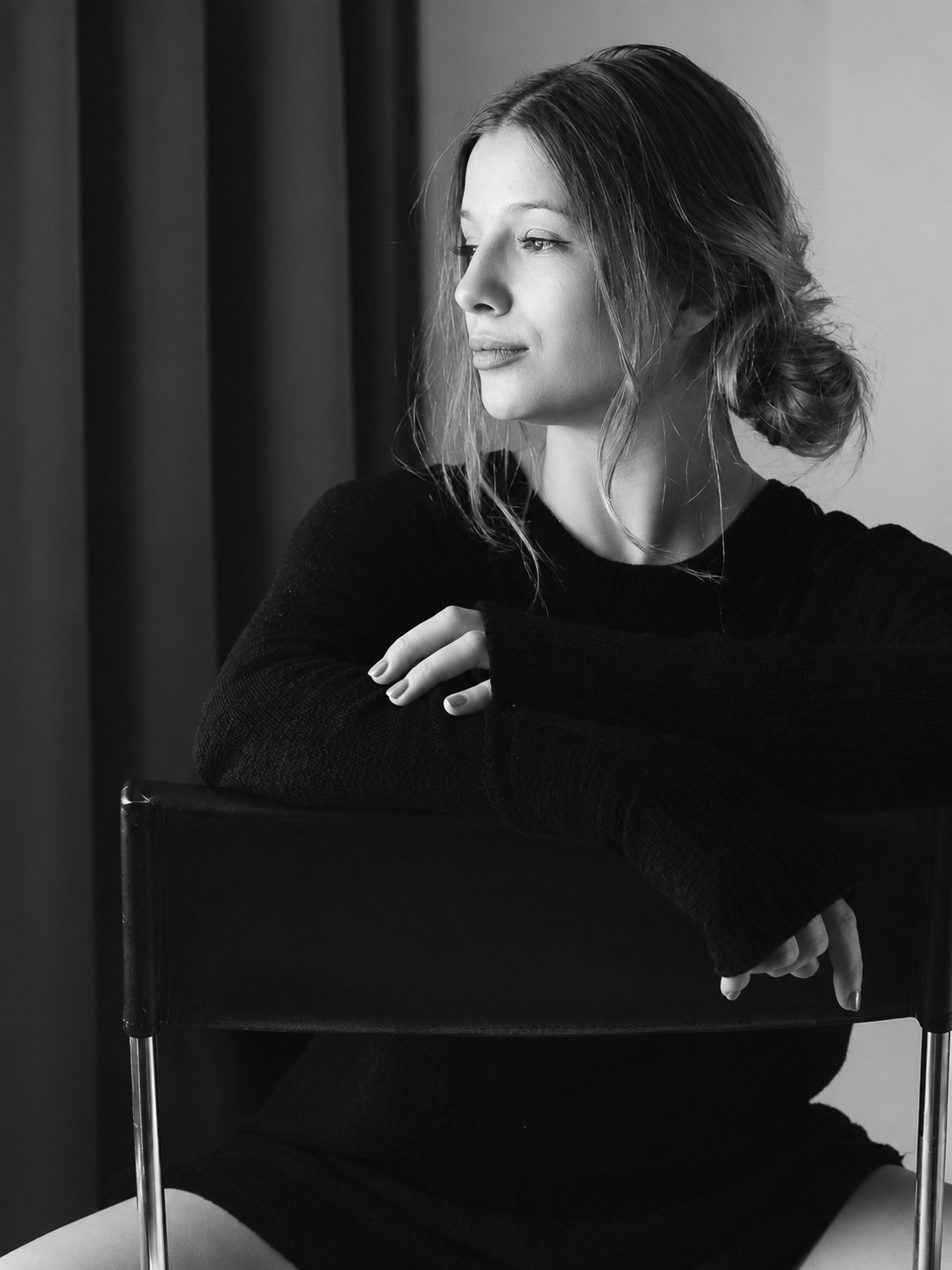Black-and-white portrait of a woman seated behind a chair, looking toward the window