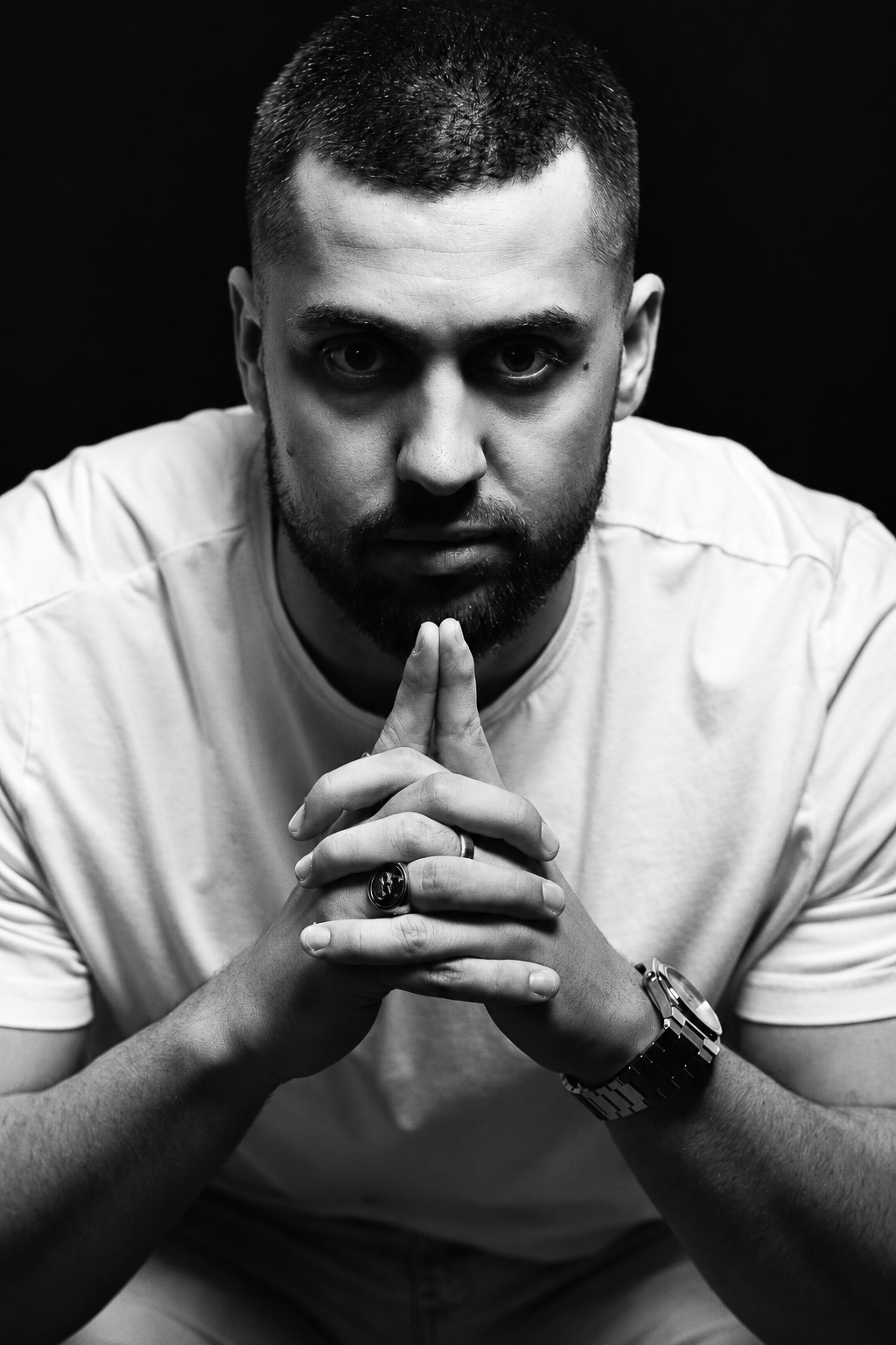 Black-and-white frontal portrait of a man in a white T-shirt with hands together